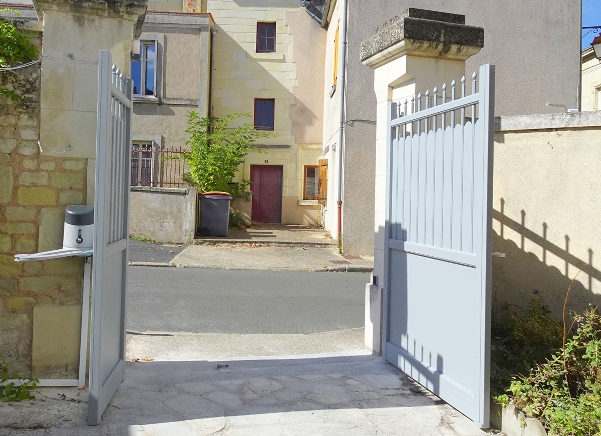 Un portail gris ouvert, encadré de piliers de pierre, donne sur une cour pavée et un vieux bâtiment à la porte rouge foncé.
