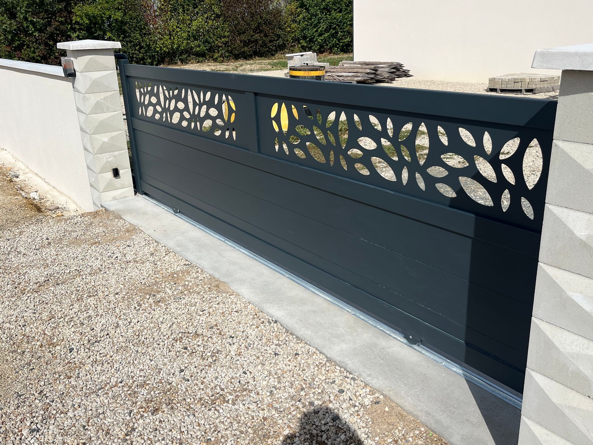 Un portail coulissant moderne gris foncé pour allée de garage, orné d'un panneau décoratif à motif de feuilles découpé au laser entre des piliers en pierre.