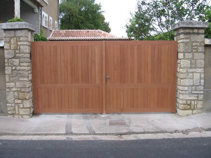Un large portail d'allée en bois brun clair, placé entre deux piliers de pierre rustiques, sur une rue pavée.