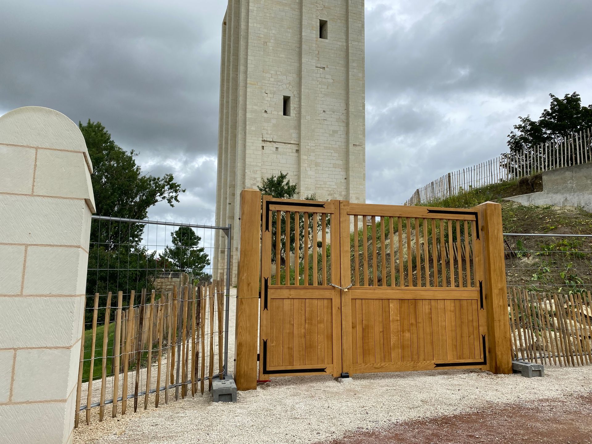Une haute tour en pierre claire se dresse derrière un portail en bois et une clôture rustique en branchages, sur un chemin de gravier sous un ciel nuageux.