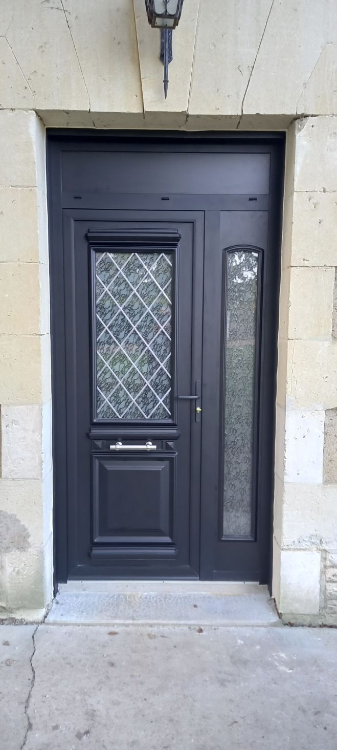 Une porte d'entrée gris foncé avec une fenêtre en verre plombé, une poignée argentée et une fente à lettres, encastrée dans un mur en pierre.