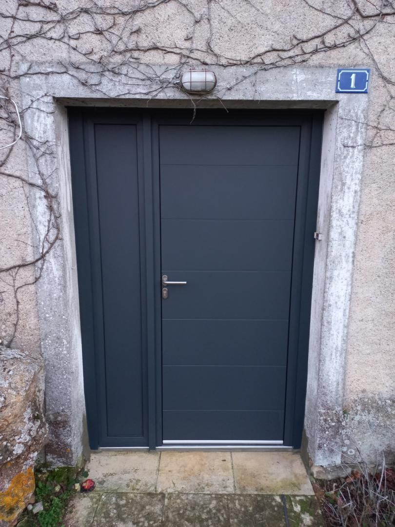 Une porte d'entrée moderne gris foncé avec un panneau latéral, installée dans un ancien cadre en pierre sur un mur recouvert de vigne nue.