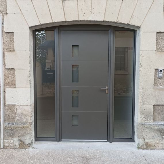 Une porte d'entrée grise et moderne, composée de quatre panneaux verticaux en verre dépoli, flanquée de hautes fenêtres latérales intégrées dans une arche en pierre.