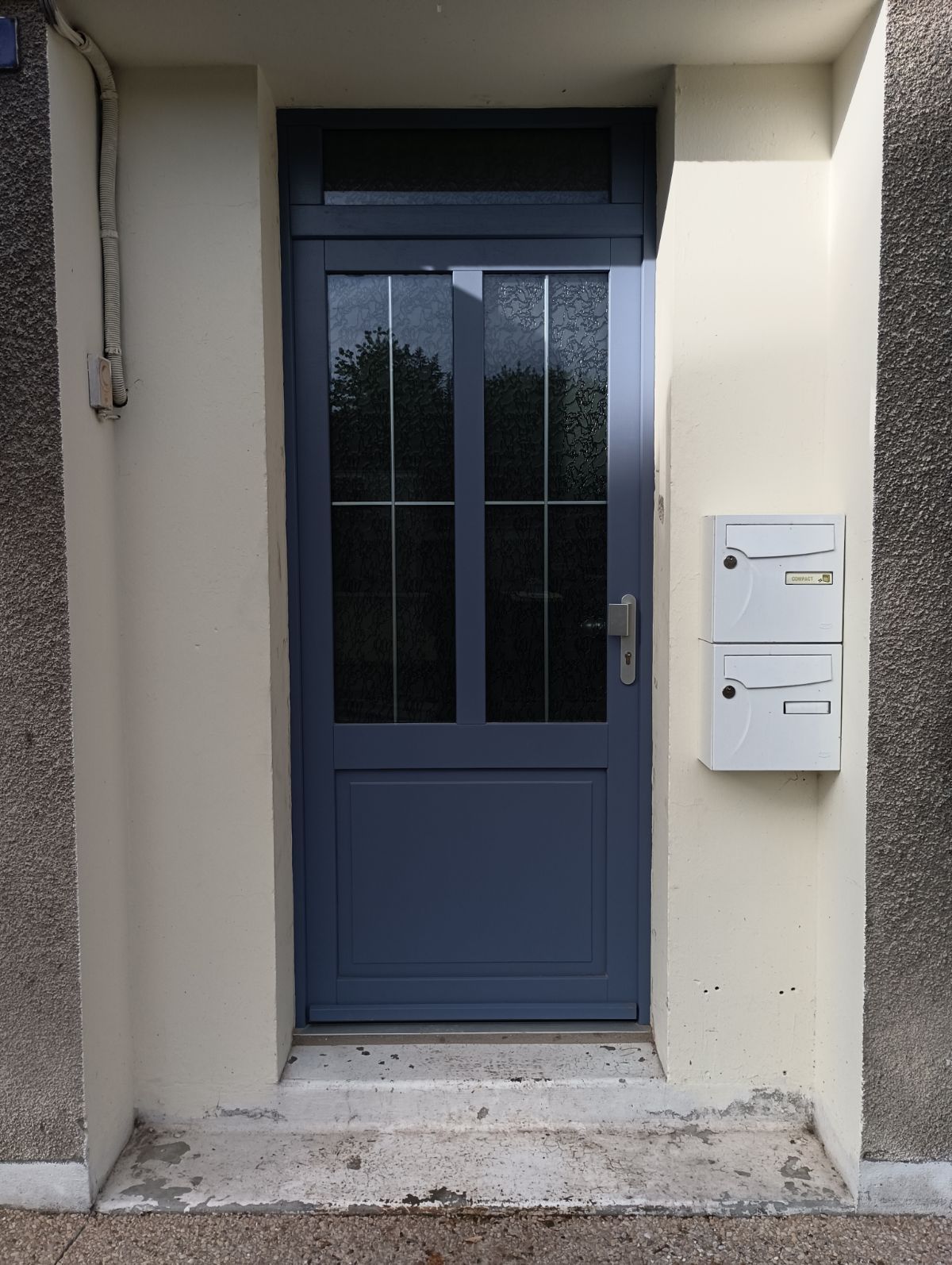 Une porte d'entrée bleu foncé à plusieurs panneaux vitrés, avec deux boîtes aux lettres blanches fixées au mur à sa droite.
