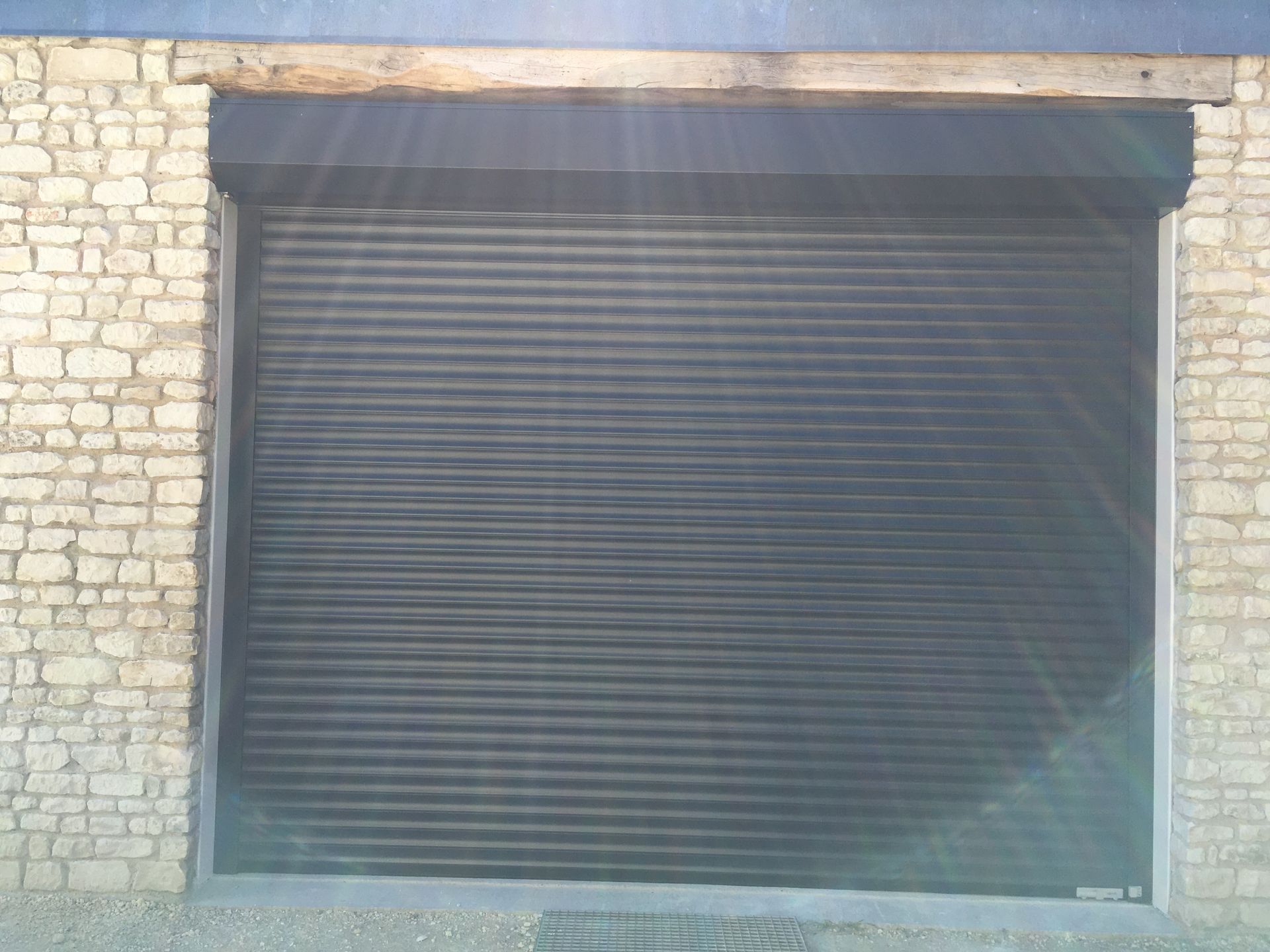 Une porte sectionnelle métallique gris foncé fermée, encastrée dans un mur de pierre sous un linteau en bois.
