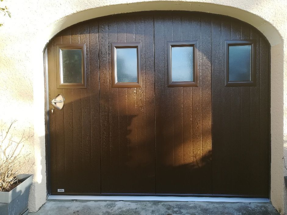 Une porte de garage à panneaux verticaux brun foncé avec quatre fenêtres rectangulaires, intégrée dans une entrée cintrée de couleur claire.