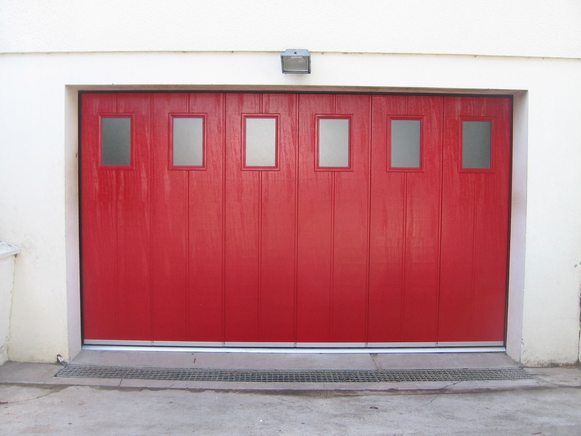 Une porte de garage rouge vif à plusieurs panneaux, avec six fenêtres rectangulaires dépolies, encastrée dans un mur extérieur blanc.