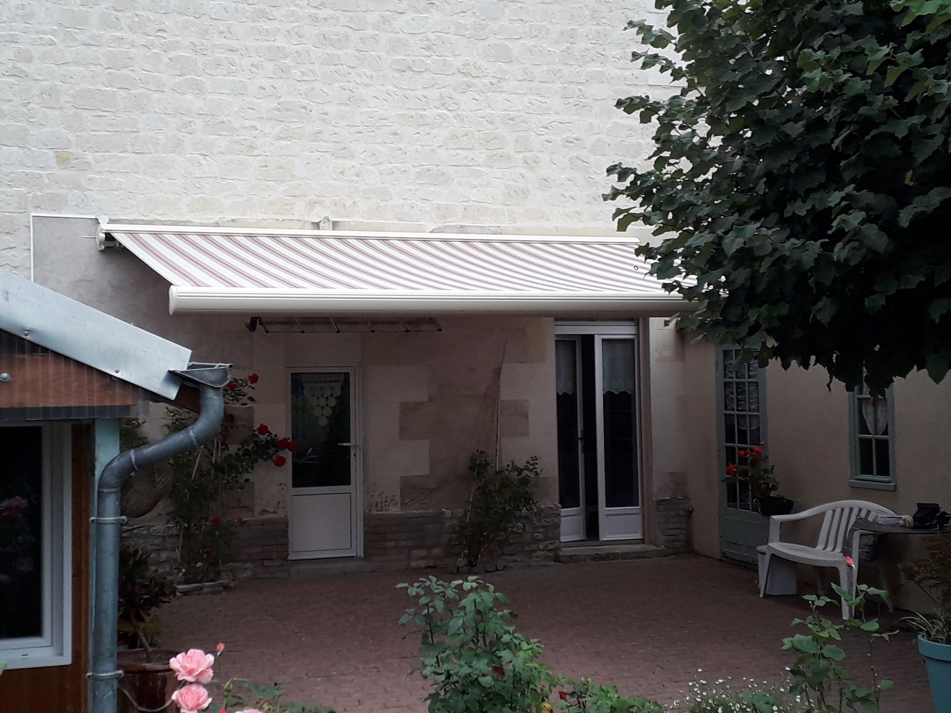 Une cour intérieure avec un store rayé au-dessus de l'entrée, des murs en pierre claire, un banc et un grand arbre.