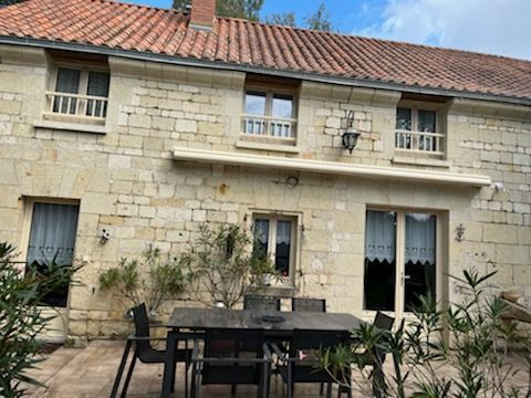 Une ferme en pierre avec un toit de tuiles, une table et des chaises sur la terrasse, et des arbres au premier plan.