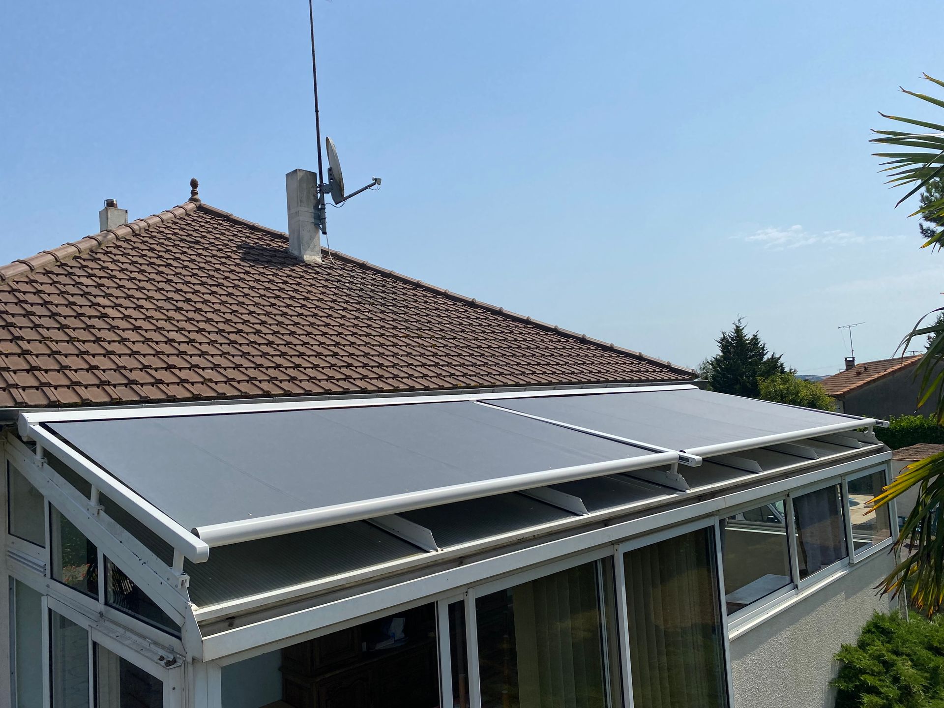 Une véranda blanche, extension de la maison, dotée de stores rétractables en tissu gris au-dessus d'un toit en verre incliné.