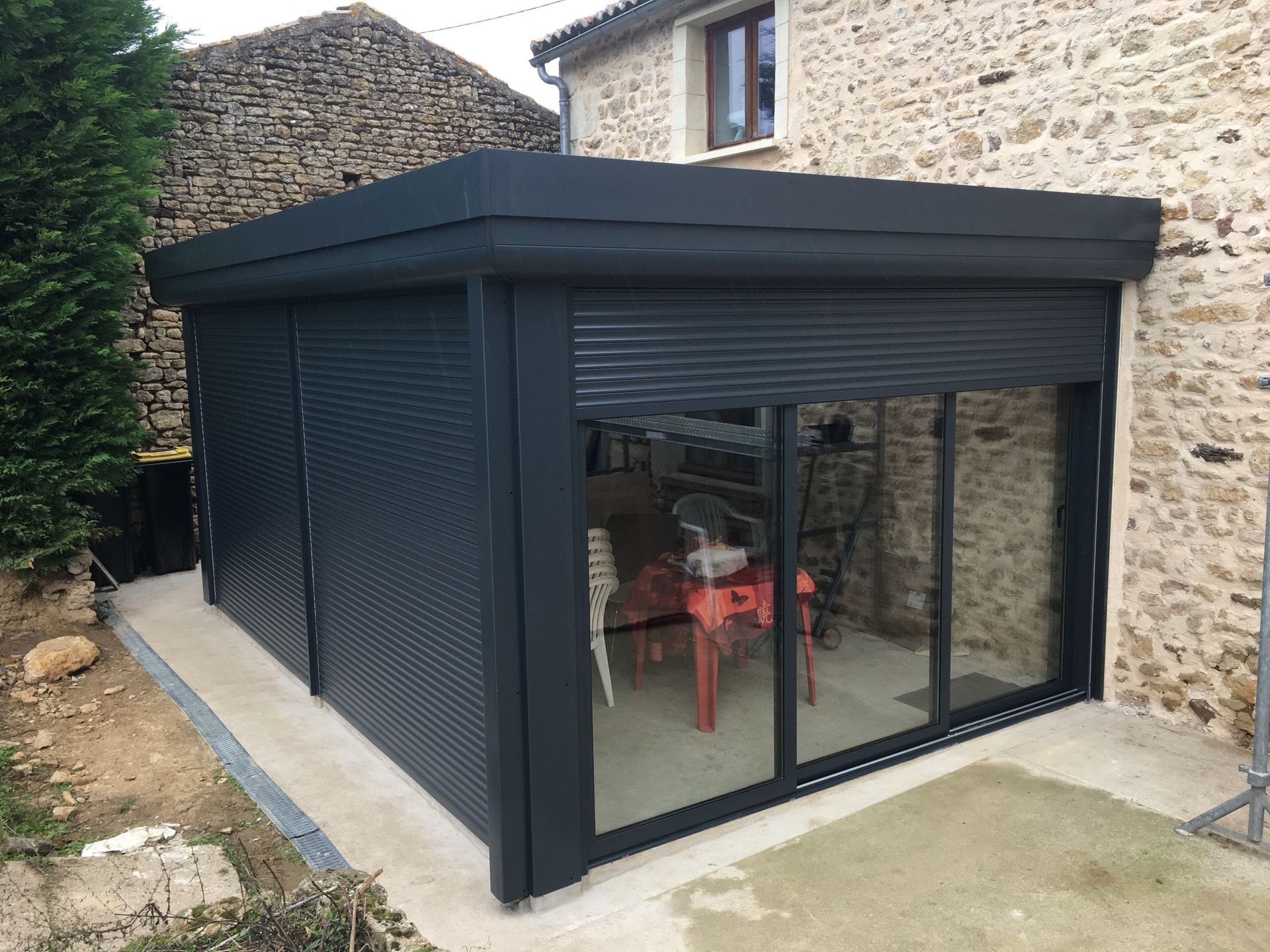 Une véranda moderne noire à toit plat, avec portes coulissantes en verre et volets de sécurité intégrés, adossée à un mur en pierre.