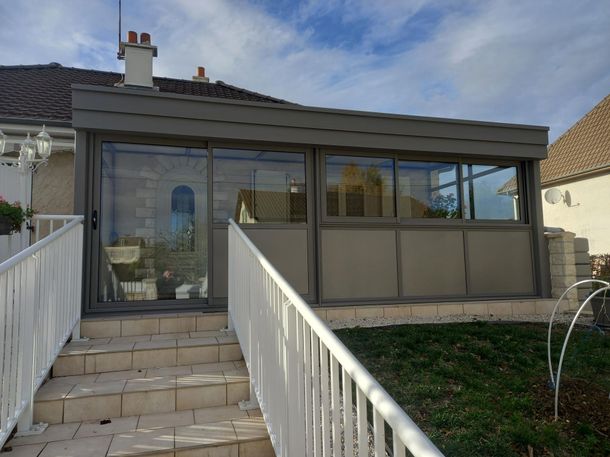 Un escalier en pierre avec une rampe en métal blanc mène à une véranda grise vitrée, attenante à une maison d'habitation beige.