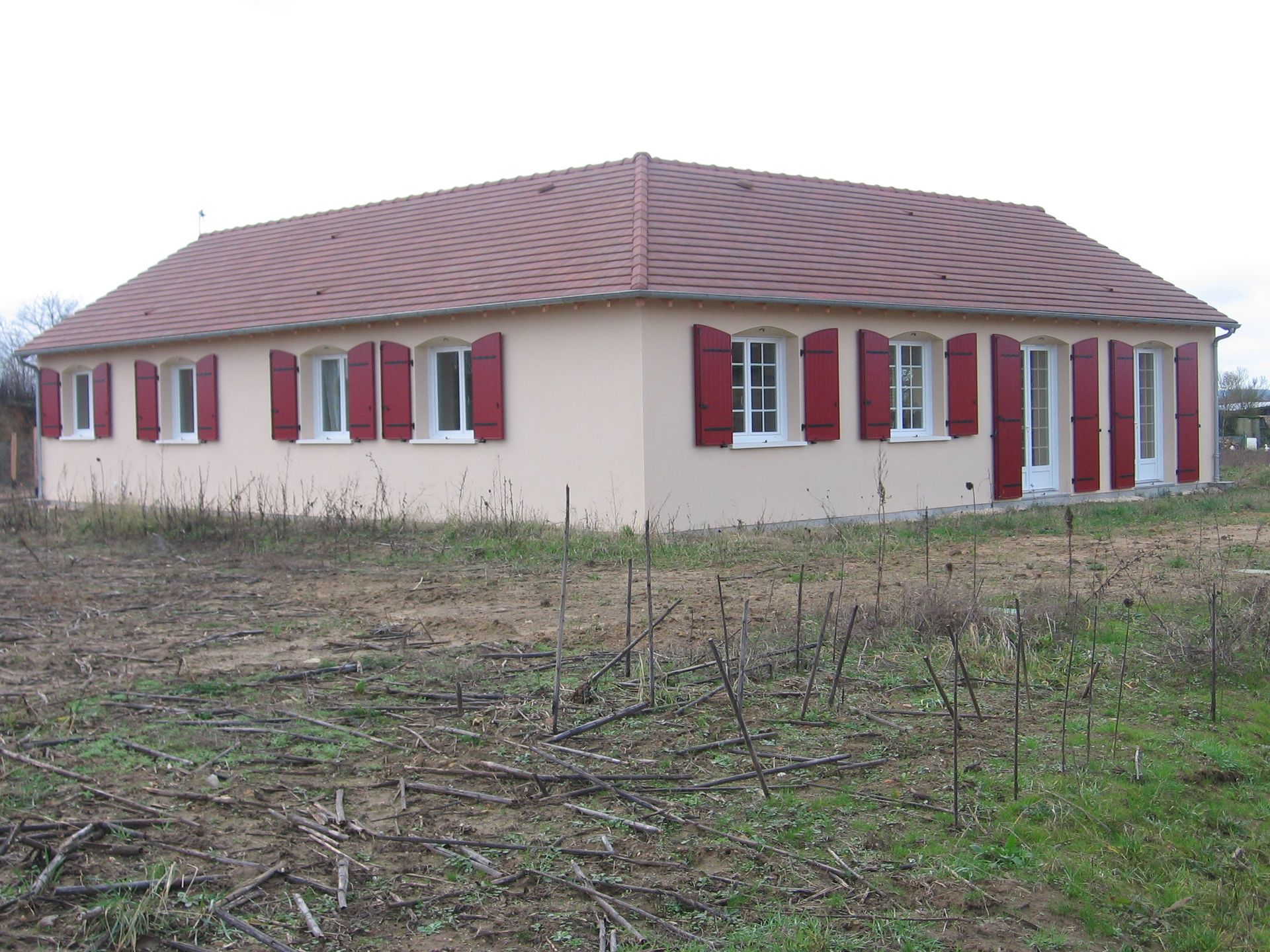 Une maison de plain-pied, rose pâle, avec un toit à quatre pans marron et des volets rouges, située dans un champ.