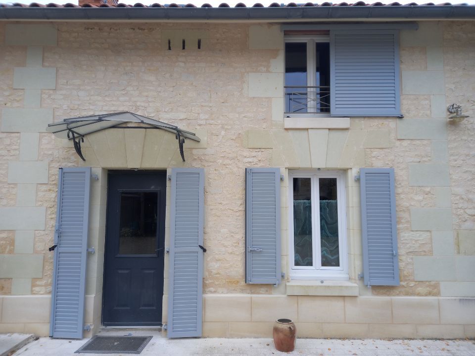 Façade en pierre d'une maison avec une porte d'entrée sombre surmontée d'un auvent métallique, une fenêtre et des volets bleus sur fond blanc.