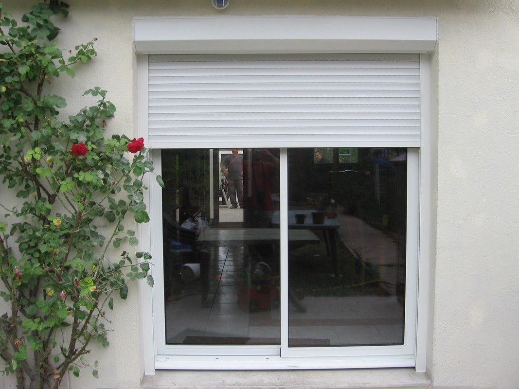 Une porte coulissante en verre avec un cadre blanc et un volet roulant blanc partiellement abaissé, adossée à un mur en stuc clair.