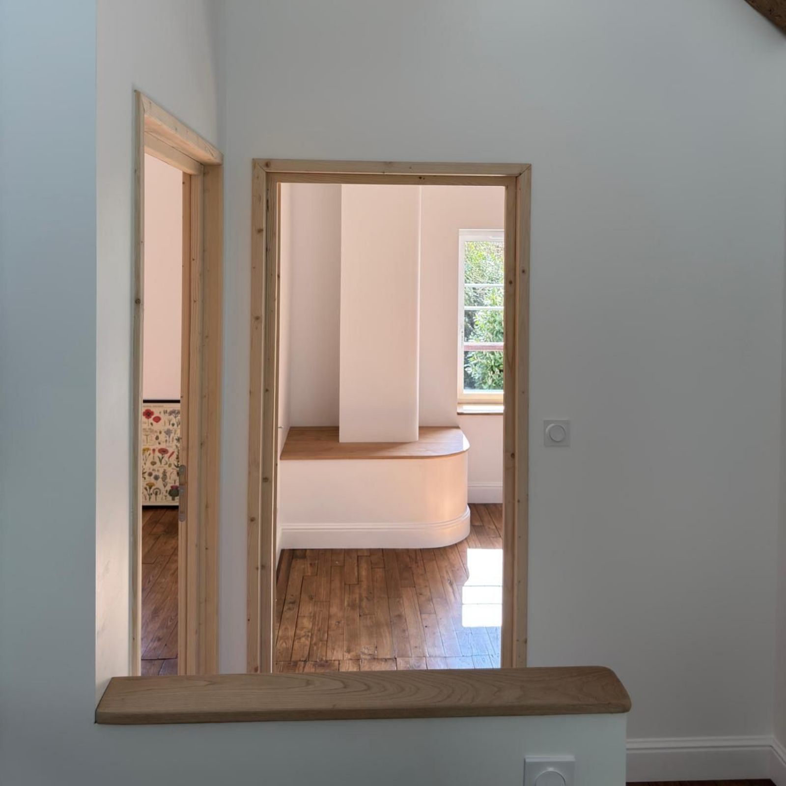 Vue à travers une porte sur une chambre lumineuse et vide, avec un parquet et une banquette blanche intégrée.