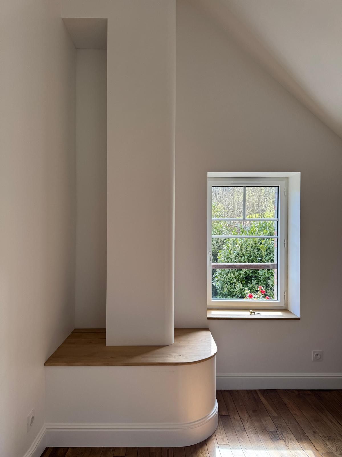Pièce blanche vide avec banquette intégrée, parquet et une petite fenêtre donnant sur de la verdure.