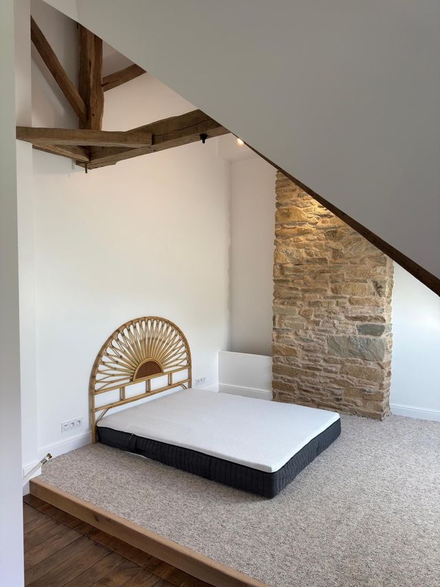 Chambre en mezzanine avec lit bas, tête de lit tressée, poutres apparentes et mur en pierre.