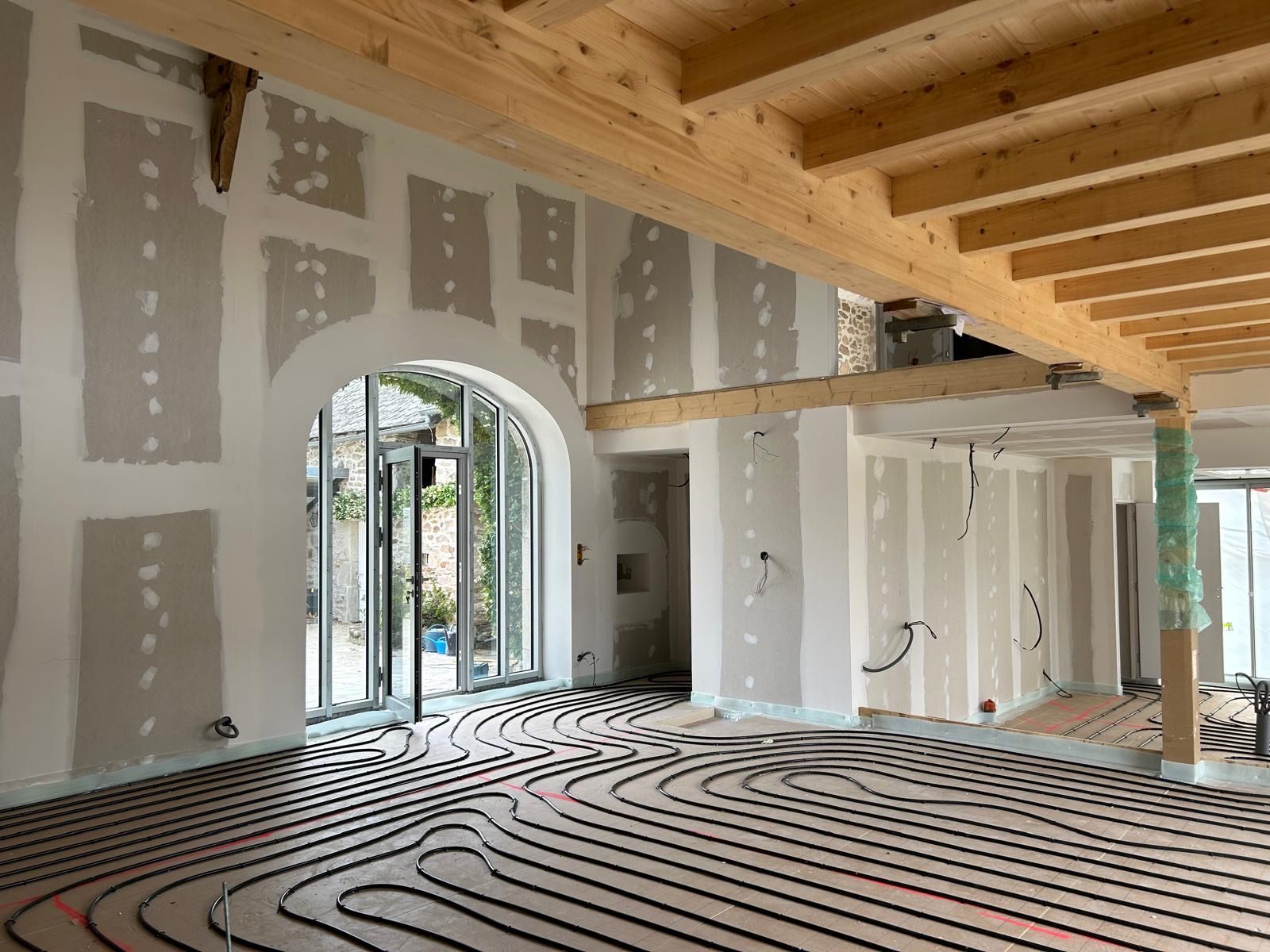 Pièce inachevée avec cloisons sèches, porte cintrée, poutres apparentes et tuyauterie de chauffage au sol.