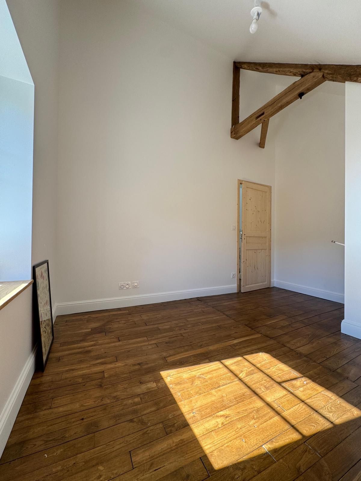 Pièce blanche vide avec parquet, poutre apparente et lumière du soleil inondant la pièce.
