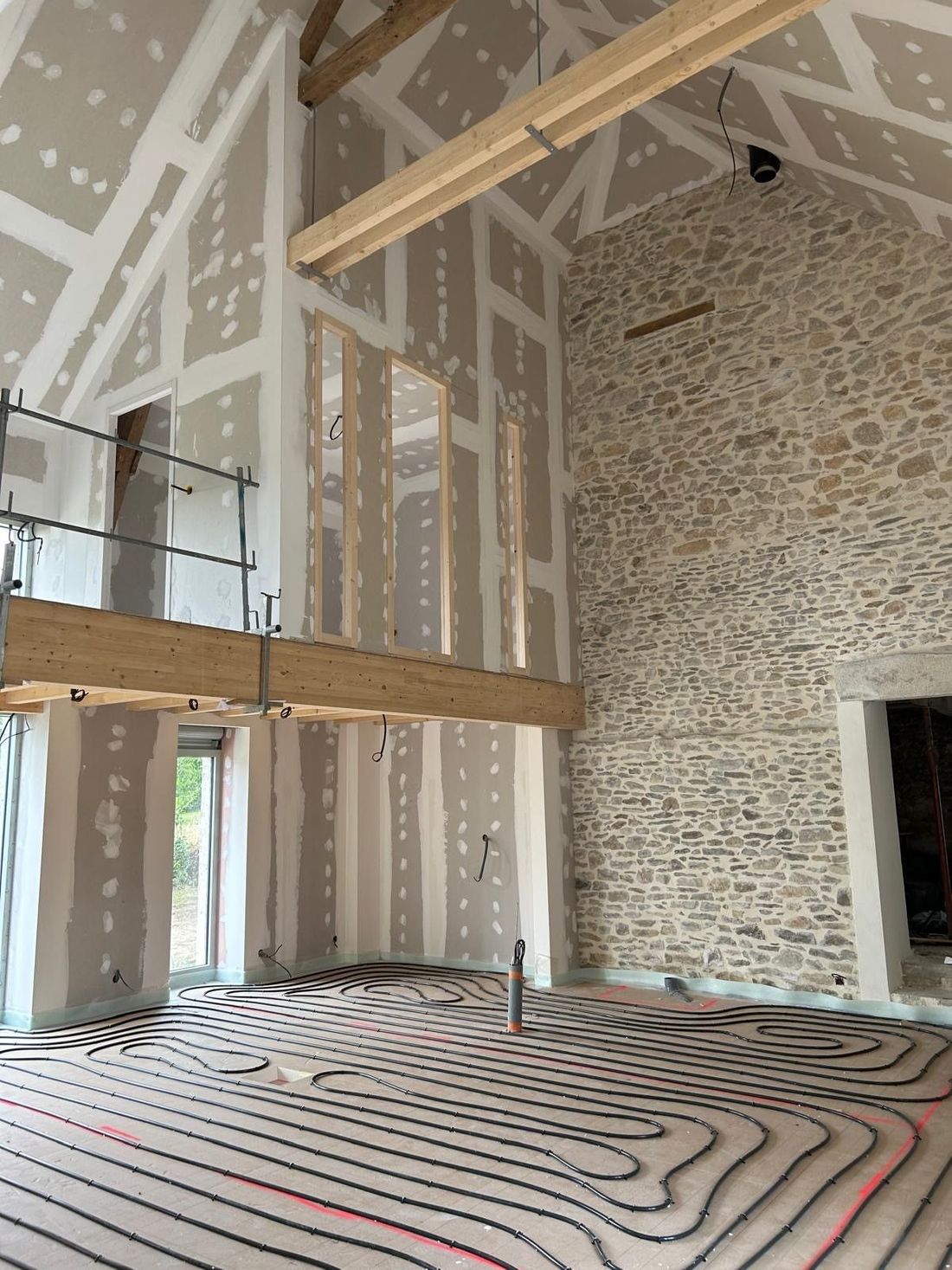Intérieur d'une pièce inachevée avec poutres apparentes, mur en pierre, mezzanine et tubes de chauffage au sol.