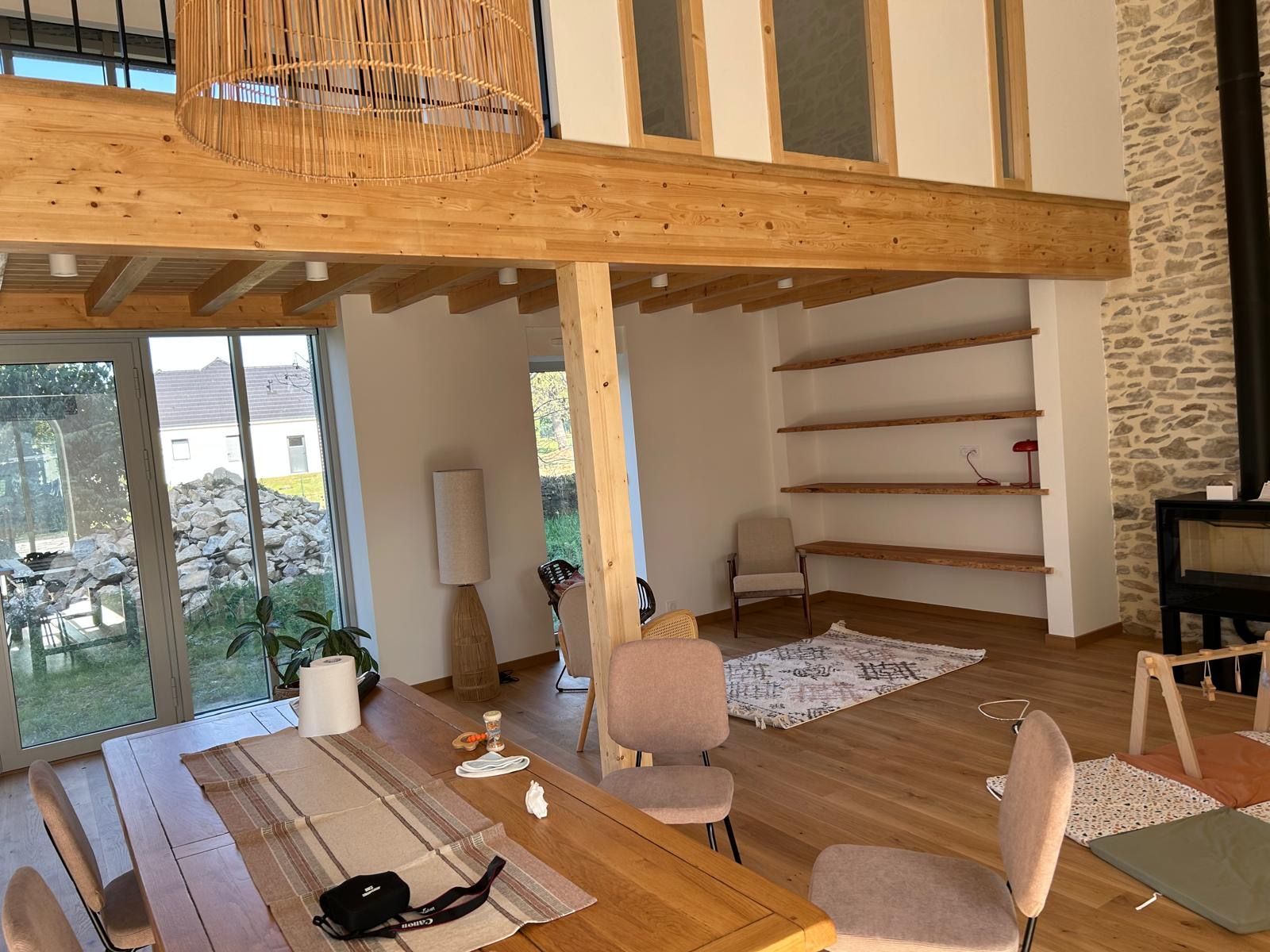 Salon moderne avec mezzanine en bois, mur en pierre, étagères et grandes portes vitrées.