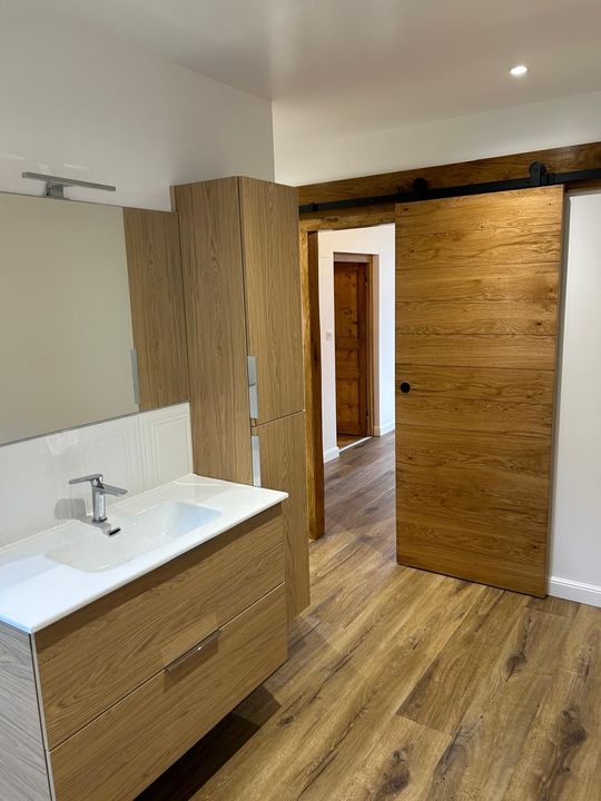 Salle de bains moderne avec meuble lavabo en bois, lavabo blanc et porte coulissante en bois donnant sur un couloir.