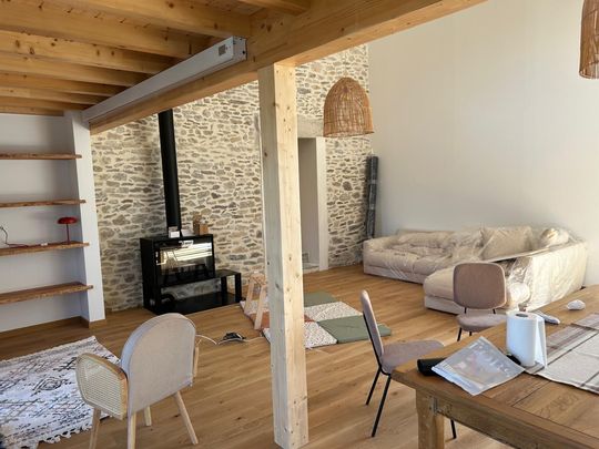 Salon rustique et chaleureux avec mur en pierre, poutres apparentes, canapé, fauteuils, étagères et petit poêle.