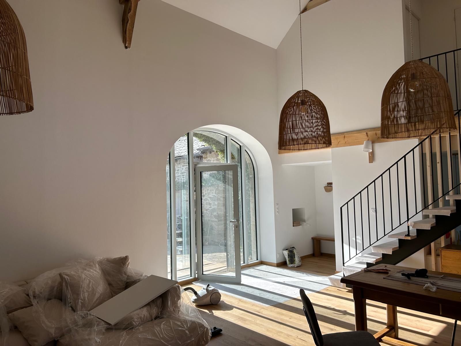 Salon lumineux avec suspensions en osier, porte vitrée cintrée, escalier et canapé beige.