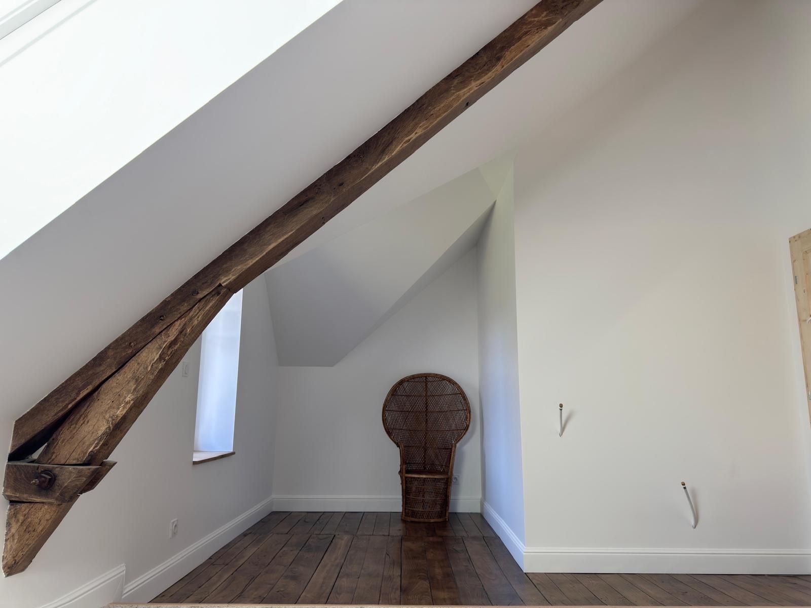 Chambre mansardée avec poutres apparentes, murs blancs, parquet foncé et fauteuil paon en osier dans une petite alcôve.