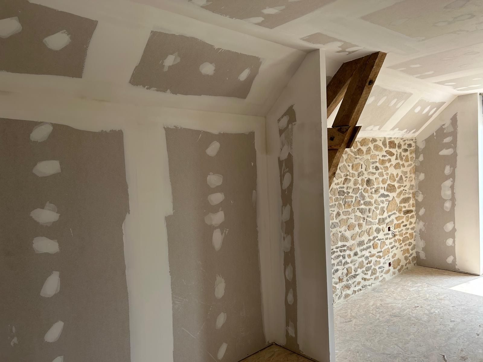 Pièce intérieure inachevée avec des plaques de plâtre enduites, des poutres apparentes et un mur en pierre.