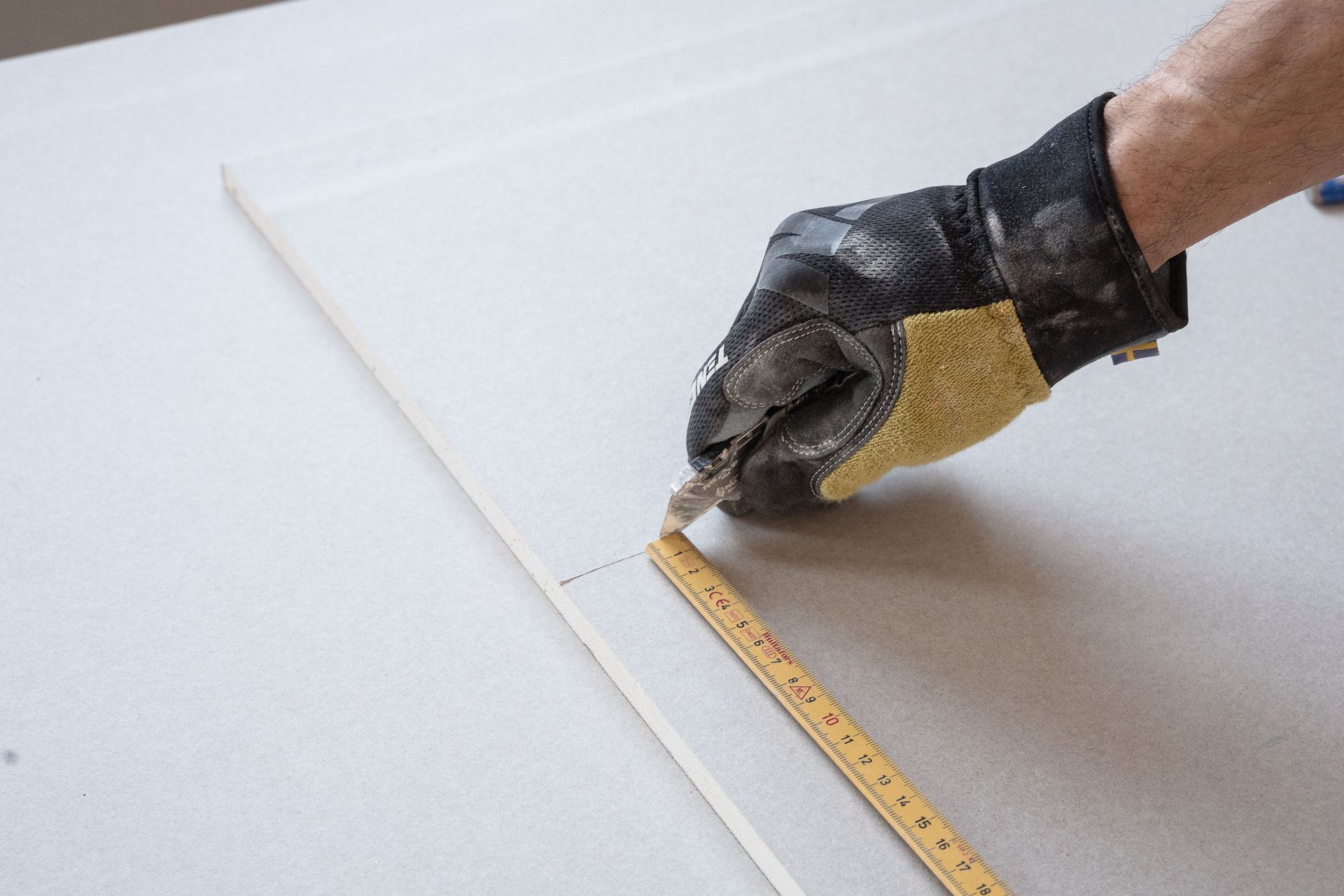 Un ouvrier mesure une plaque de placo tout en la découpant avec un couteau de précision.