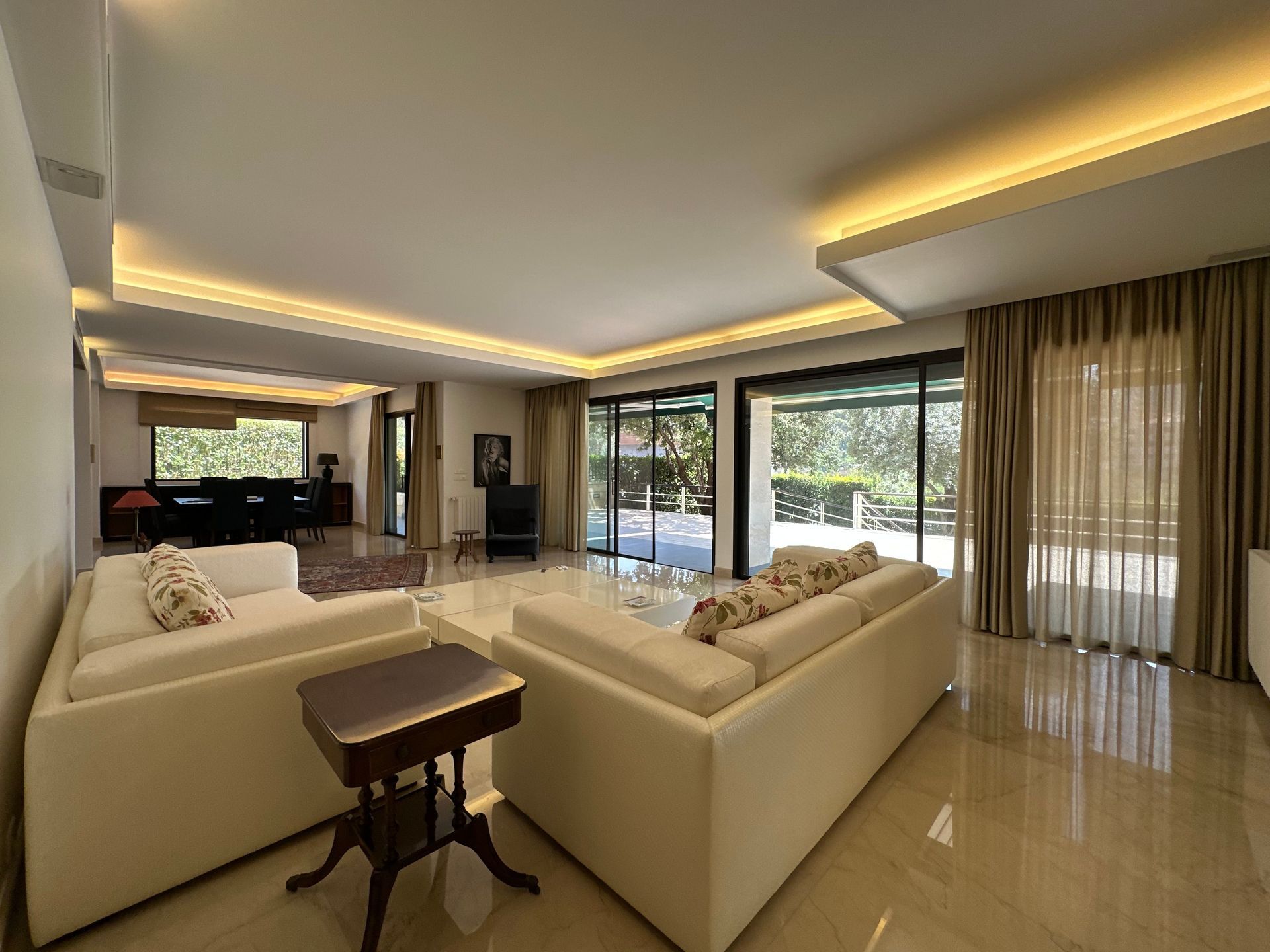 Salon moderne avec canapés beiges, carrelage brillant et grandes fenêtres donnant sur la verdure.
