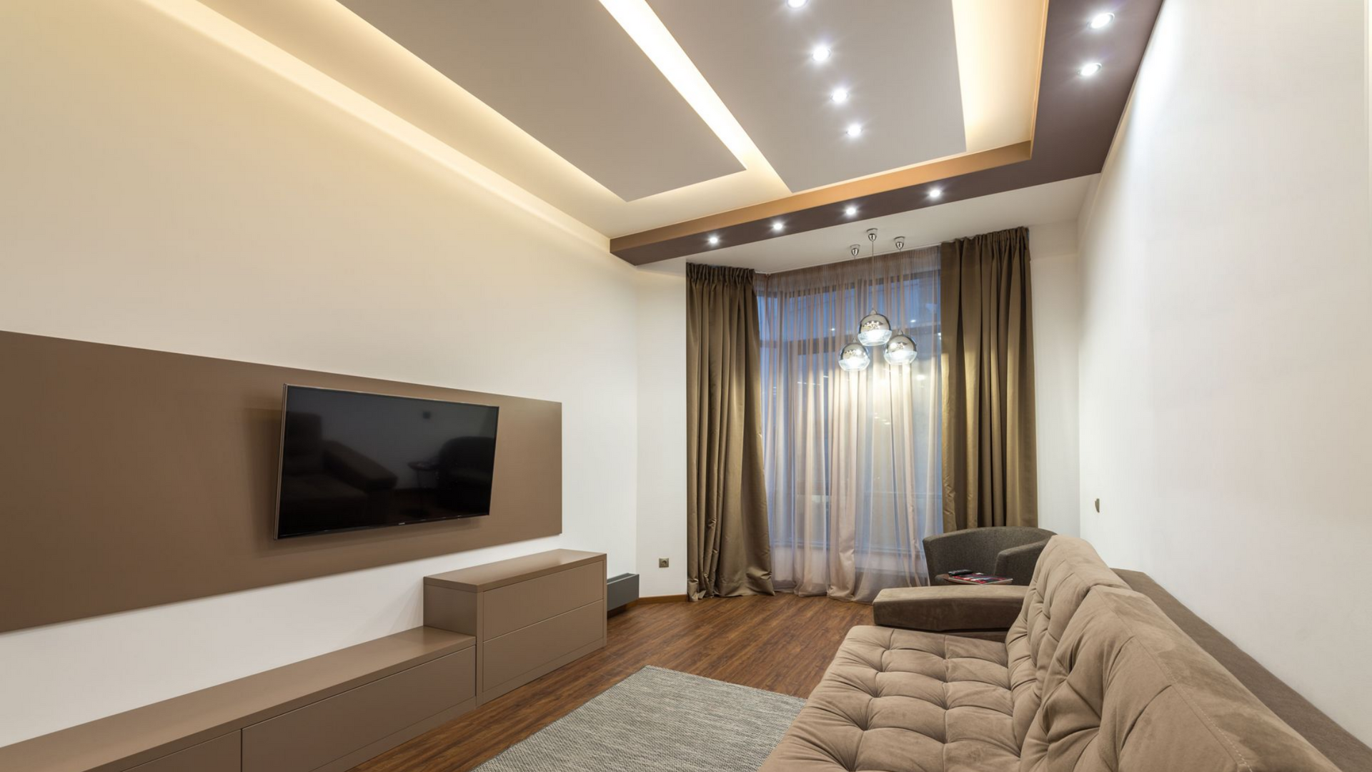 Salon moderne avec canapé beige, télévision murale et éclairage chaleureux au plafond.