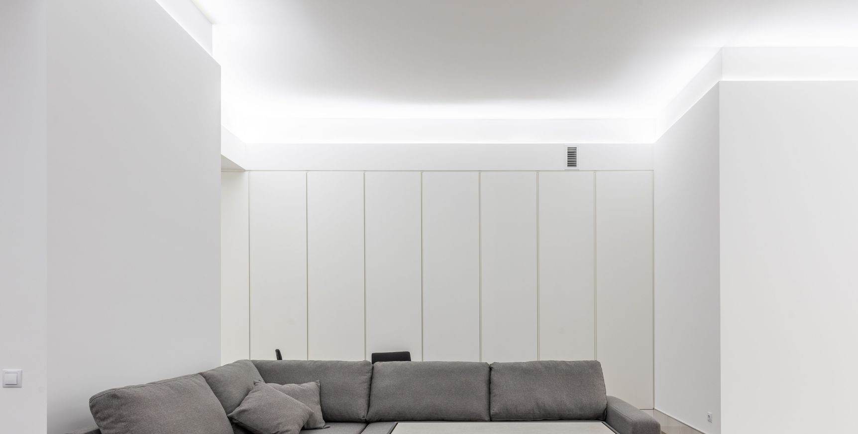 Salon minimaliste blanc avec un canapé d'angle gris et un éclairage LED au plafond.