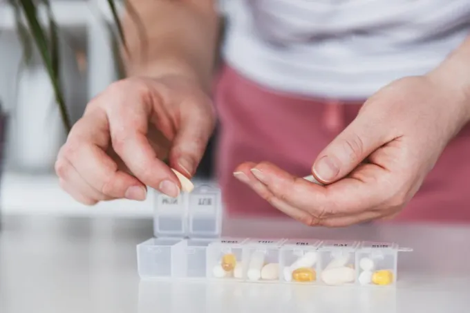 Manos tomando pastillas de un organizador de medicamentos diario que está sobre una mesa.