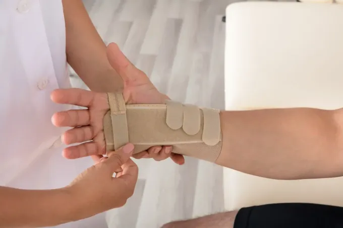 Un profesional sanitario ajusta una muñequera beige en la mano de un paciente en un entorno clínico.