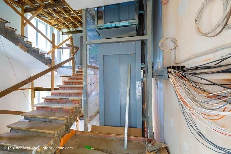 Aufzugsschacht neben einem im Bau befindlichen Treppenhaus; Kabel, grauer Aufzug und Holzbalken sind zu sehen.