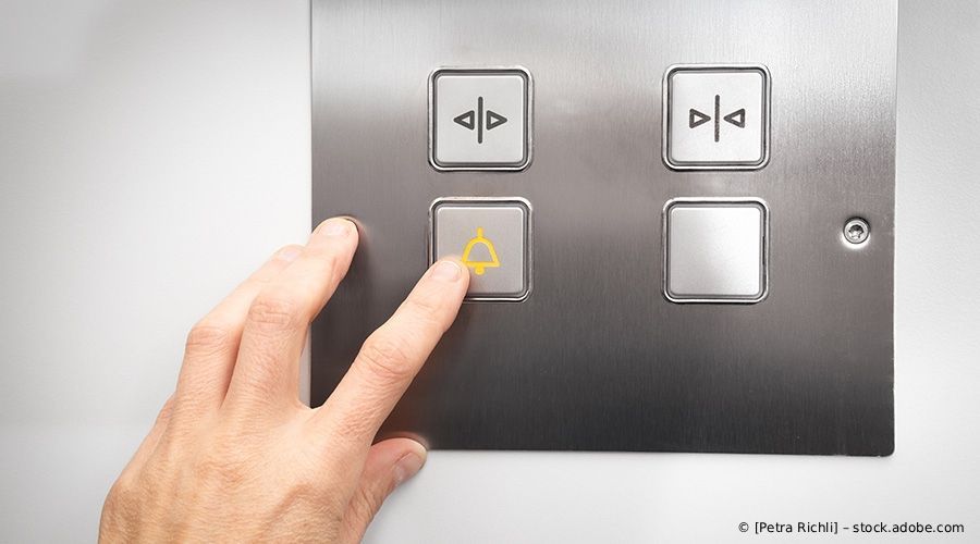 Eine Hand drückt den Aufzugsrufknopf mit einem Glockensymbol auf einer Edelstahlplatte.