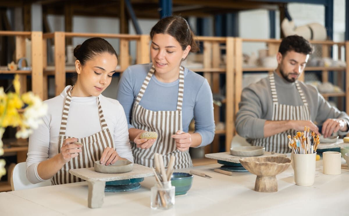 Tres personas haciendo cerámica en una mesa de taller, moldeando arcilla con herramientas y delantales puestos.
