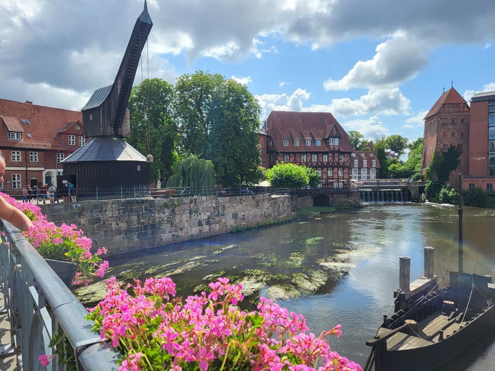 Holzkran, Fluss, Gebäude und rosa Blumen unter blauem Himmel.