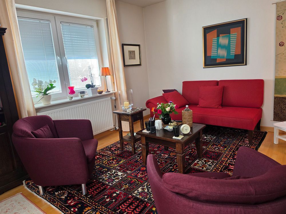Gemütliches Wohnzimmer mit roten und violetten Sesseln, gemustertem Teppich, Couchtisch und Fensterpflanzen