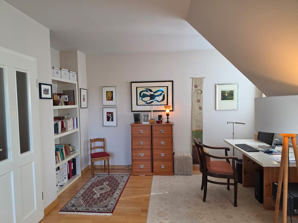 Helles Heimbüro mit Schreibtisch, Stuhl, Bücherregalen, Kommode, Teppich und Dachschräge.
