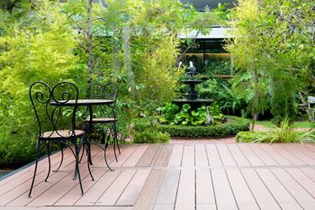 Holzfläche mit Gartenstühlen und Tisch, im Hintergrund ein Garten mit Springbrunnen