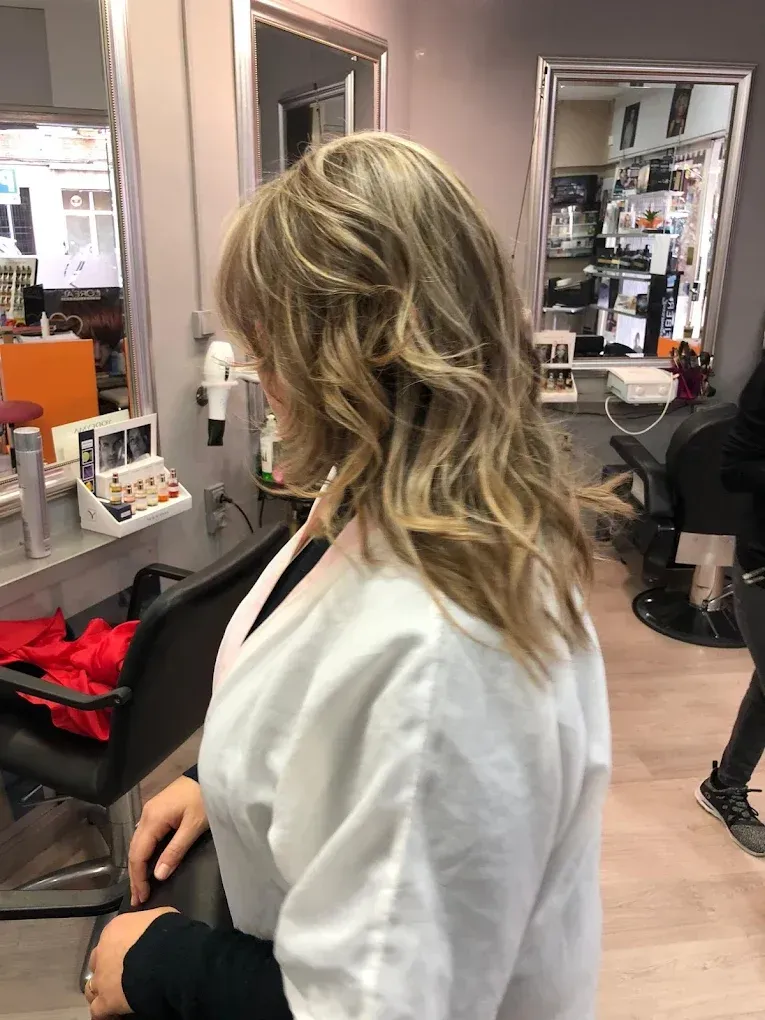 Mujer con cabello rubio ondulado sentada en una silla de peluquería, frente a un espejo en un salón de belleza.
