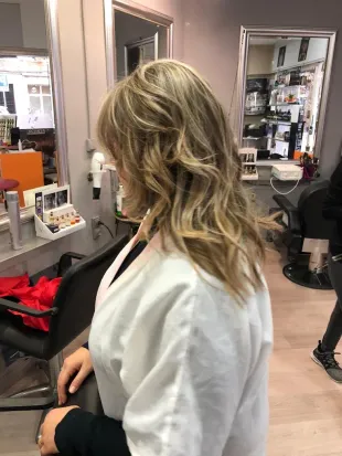 Mujer con cabello rubio ondulado sentada en una silla de peluquería, frente a un espejo en un salón de belleza.