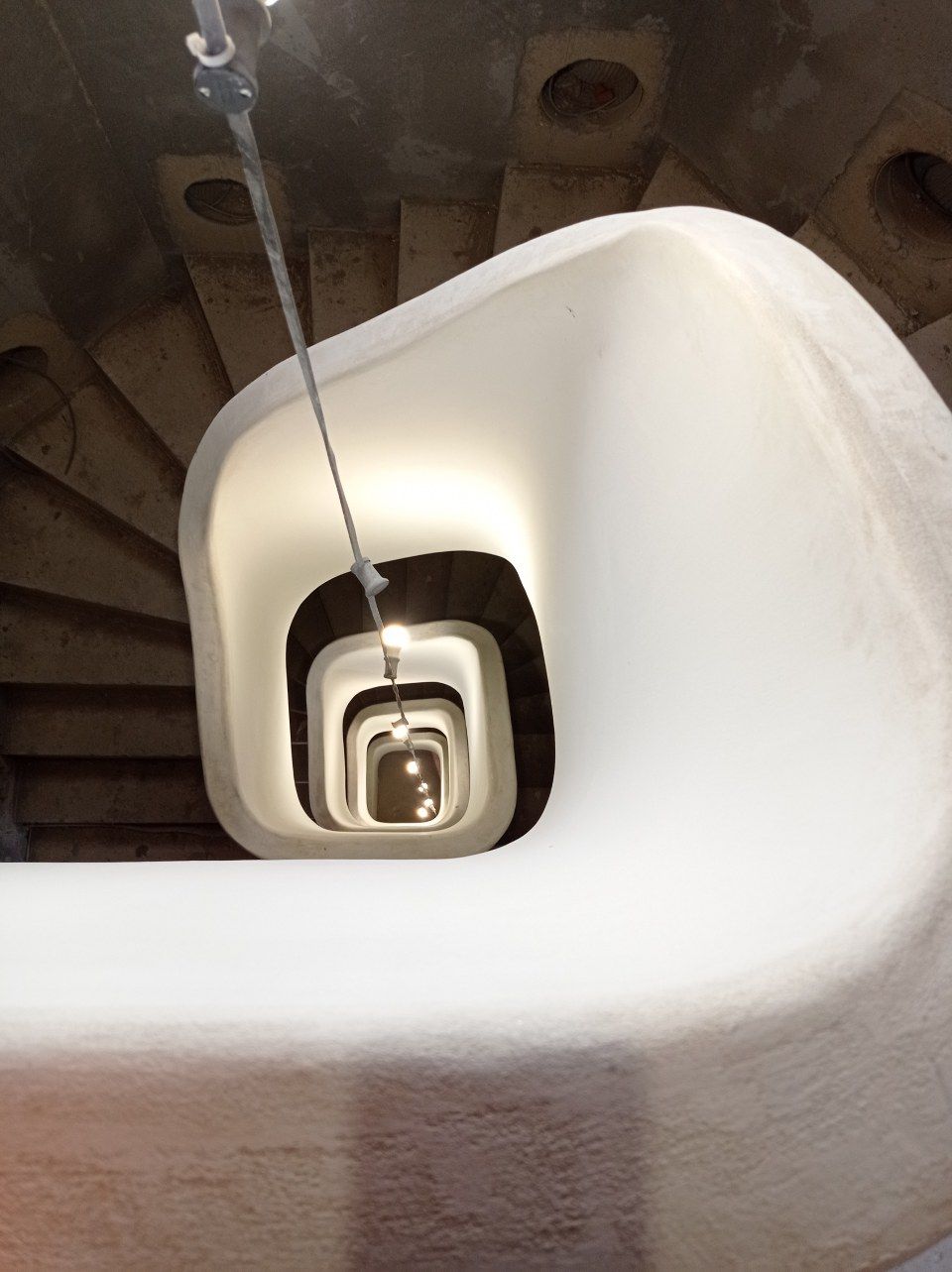 Blick hinunter auf eine Wendeltreppe mit einer zentralen Hängelampe, die einen sich wiederholenden Tunneleffekt erzeugt.