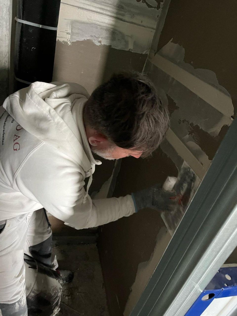 Eine Person in einem weissen Kapuzenpullover trägt bei einer Hausrenovierung mit einer Kelle Gips an einer Wand auf.