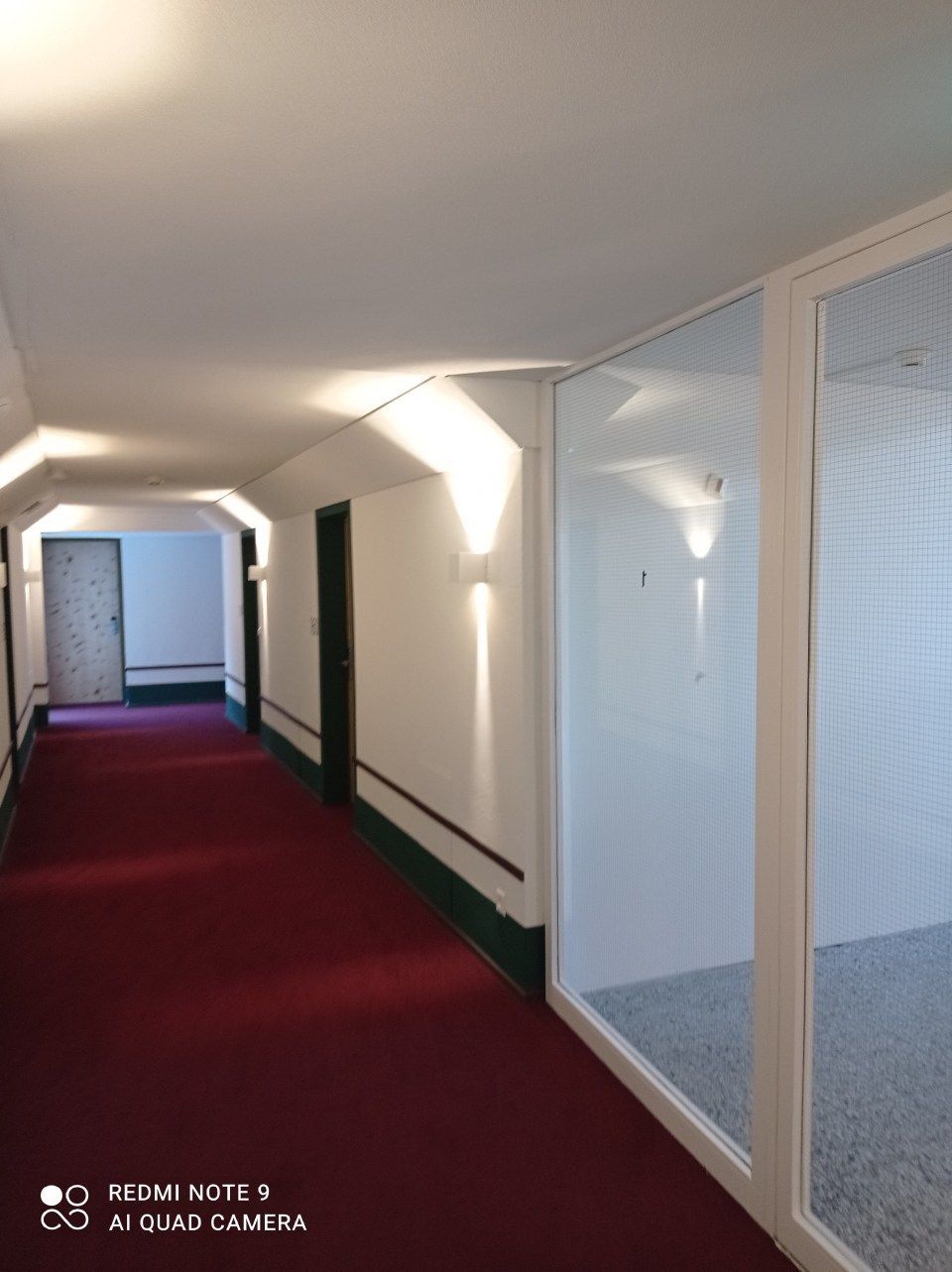 Ein langer, überdachter Flur mit rotem Teppichboden, weissen Wänden, dunklen Türen & rechts davon verglasten Bürotrennwänden.