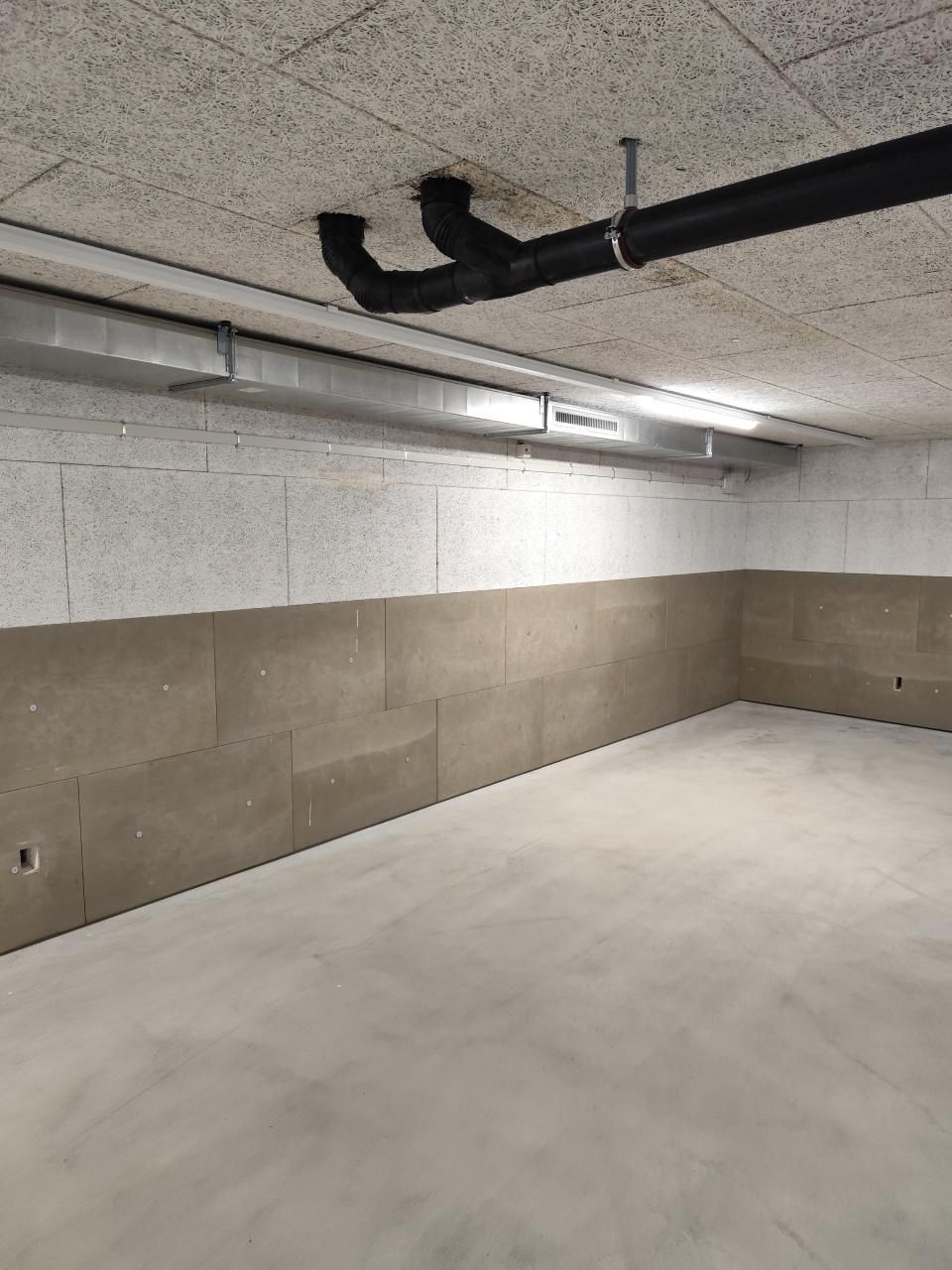 Ein unfertiger Kellerraum mit Betonsteinwänden, grauem Betonboden und freiliegenden Deckenrohren und -kanälen.